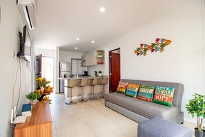 Ein modernes Wohnzimmer mit einem grauen Sofa, einem Holztisch und bunten Kissen. Im Hintergrund ist eine Küche mit Barhocker erkennbar.