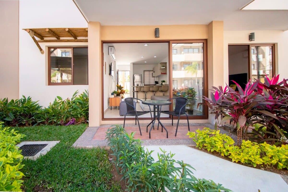 A modern outdoor patio with a dining area, a small kitchen visible through glass doors, and a variety of plants.