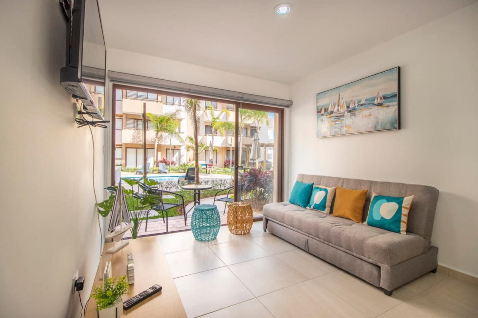 Ein modernes Wohnzimmer mit einem grauen Sofa, einem Gemälde von Segelbooten und Blick auf einen Pool und Gebäude im Freien.