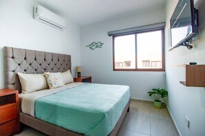 Ein Schlafzimmer mit einem gesteppten Kopfteil, einem Bett mit grüner Decke, einem Nachttisch mit Lampe, einem Fenster mit Jalousien und einem an der Wand montierten Fernseher.