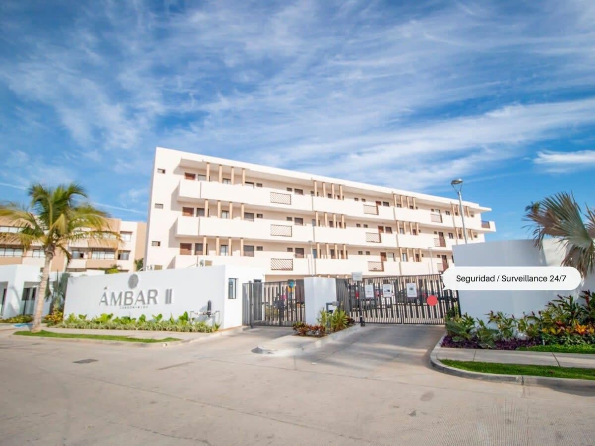 Un hotel a più piani con un ingresso chiuso da cancello, un cartello che indica sicurezza attiva 24 ore su 24, e una zona verde con palme.