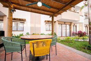 Eine Terrassengaststätte mit rundem Tisch und Stühlen, einem Deckenventilator und Blick auf ein Gebäude mit großen Fenstern.