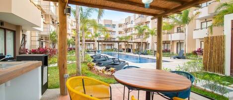 A covered outdoor area with a pool, yellow chairs, and a wooden table.
