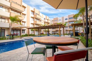 Ein Poolbereich mit Holztisch und Stühlen, umgeben von einem Pool und einem Gebäude mit Balkonen.