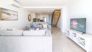A modern living room with a grey sofa, a TV on a white cabinet, and a staircase leading to an upper level. - Superb 2-Bed Apartment with Scenic Bay Views (Batemans Bay)
