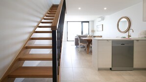A modern kitchen with a central island, a dining area with a table and chairs, and a staircase leading to another level. - Superb 2-Bed Apartment with Scenic Bay Views (Batemans Bay)