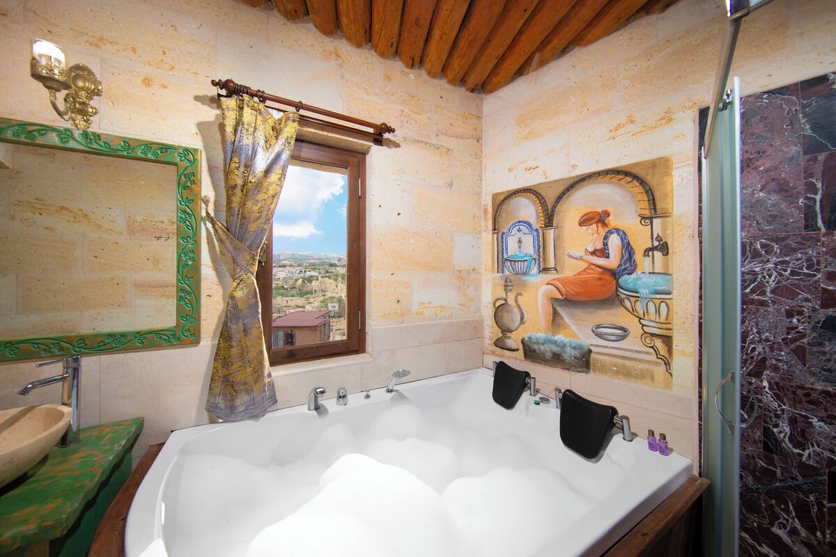 A bathroom with a large bathtub, a window with a view, a mirror with a decorative frame, and a tiled wall.