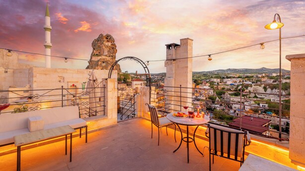 A rooftop terrace with a table and chairs, a view of a cityscape, and a prominent rock formation. - Turkan Cave Hotel (Ürgüp)