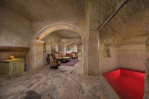 Una habitación con paredes de piedra, piscina con mosaicos rojos, muebles de madera y suelo de piedra.