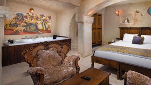 A room with a bed, a sofa, a wooden table, a bathroom with a sink, and a decorative wall painting. - Turkan Cave Hotel (Ürgüp)
