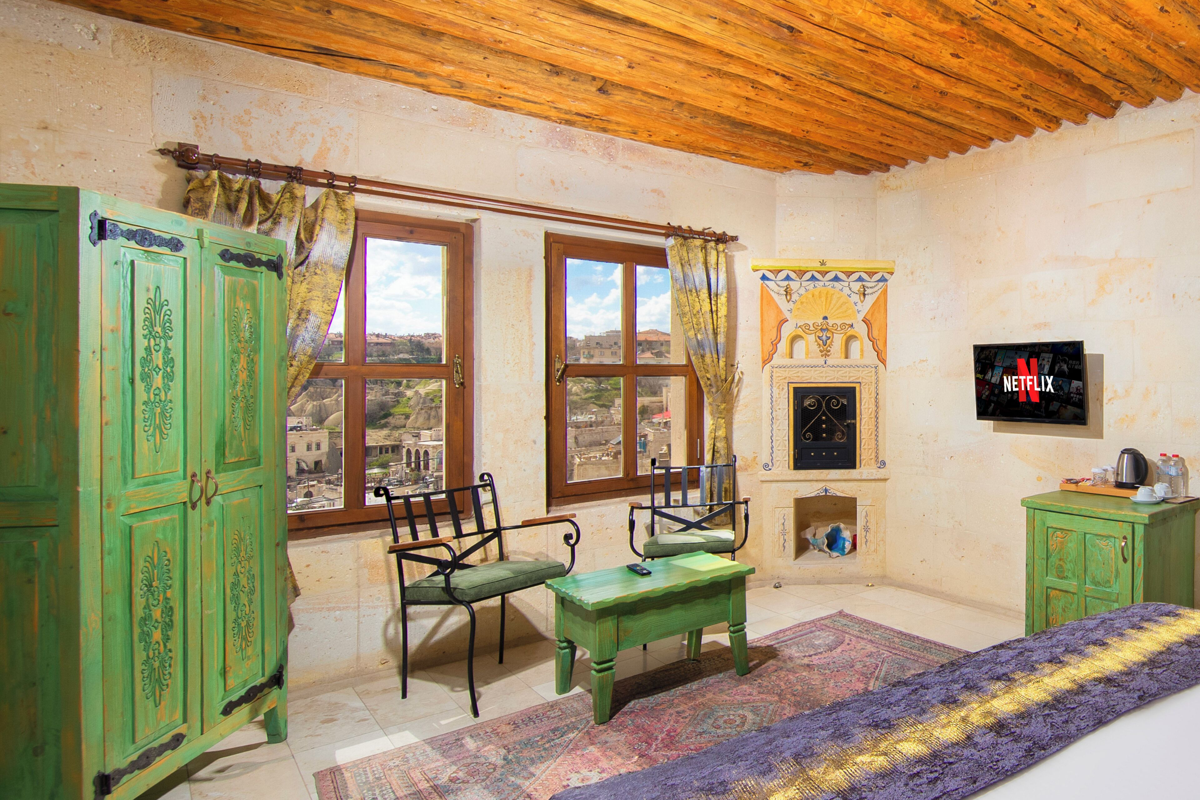 A room with a wooden ceiling, two windows with curtains, a green cabinet, a wooden table, a bench, a fireplace, and a television.