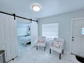 A room with white walls, a bed with floral bedding, two white chairs with floral cushions, a ceiling light, and a window with blinds. - Complete Remodel Charmin home near bush garden,hard rock casino, Moffitt,Usf!! (Tampa)