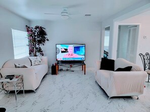 A living room with two white sofas, a TV, a ceiling fan, and a plant. - Complete Remodel Charmin home near bush garden,hard rock casino, Moffitt,Usf!! (Tampa)
