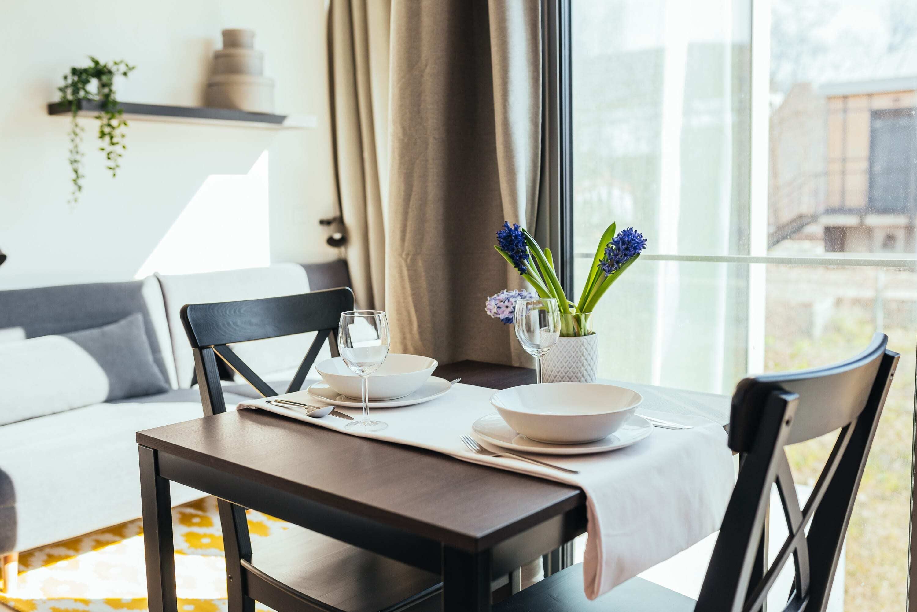 Un coin repas avec une table dressée pour deux personnes, un vase rempli de fleurs et une vue sur l’extérieur par une fenêtre.