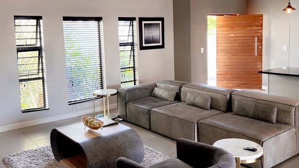 Uma sala de estar moderna com sofá seccional, mesa de centro redonda e chaise longue. Há janelas amplas com persianas, uma parede de destaque em madeira e uma luminária pendente.