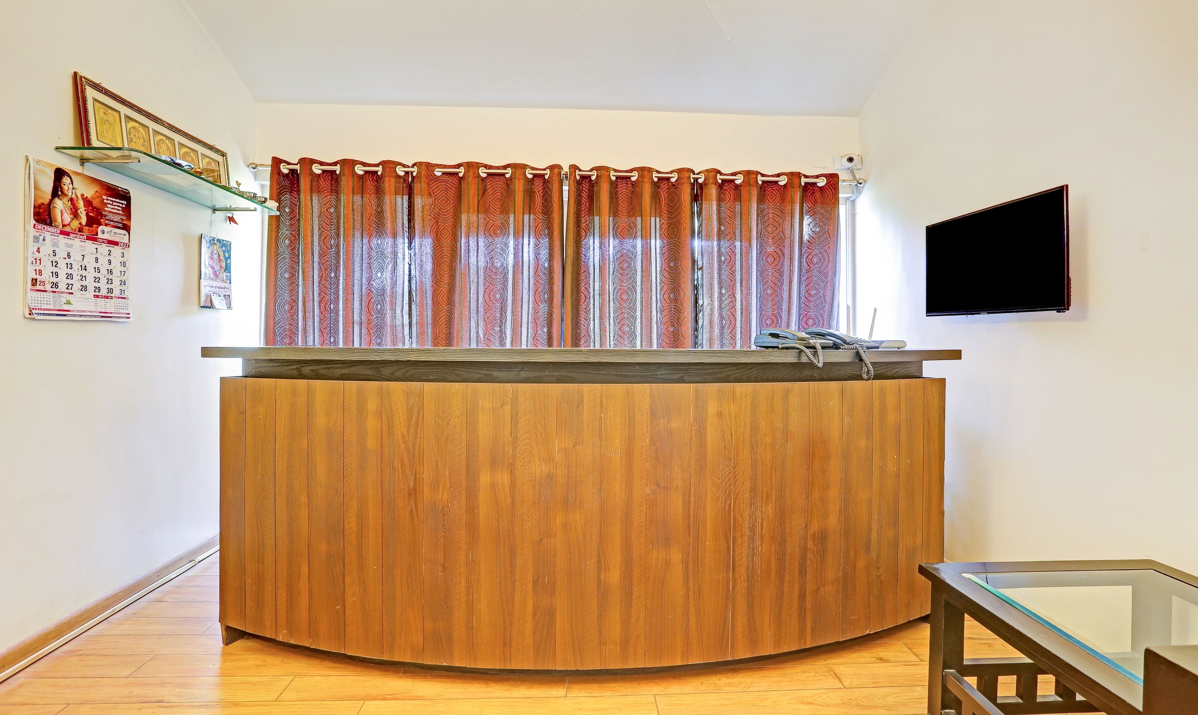 A reception desk with a wooden finish, a glass top, and a telephone. There is a calendar on the wall to the left and a flat-screen TV mounted on the wall to the right.