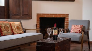 A cozy living room with a fireplace, a sofa with patterned cushions, a wooden coffee table with a bottle of wine and two glasses, and a chair with a floral pillow. - Casita del Escritor (Antigua Guatemala)