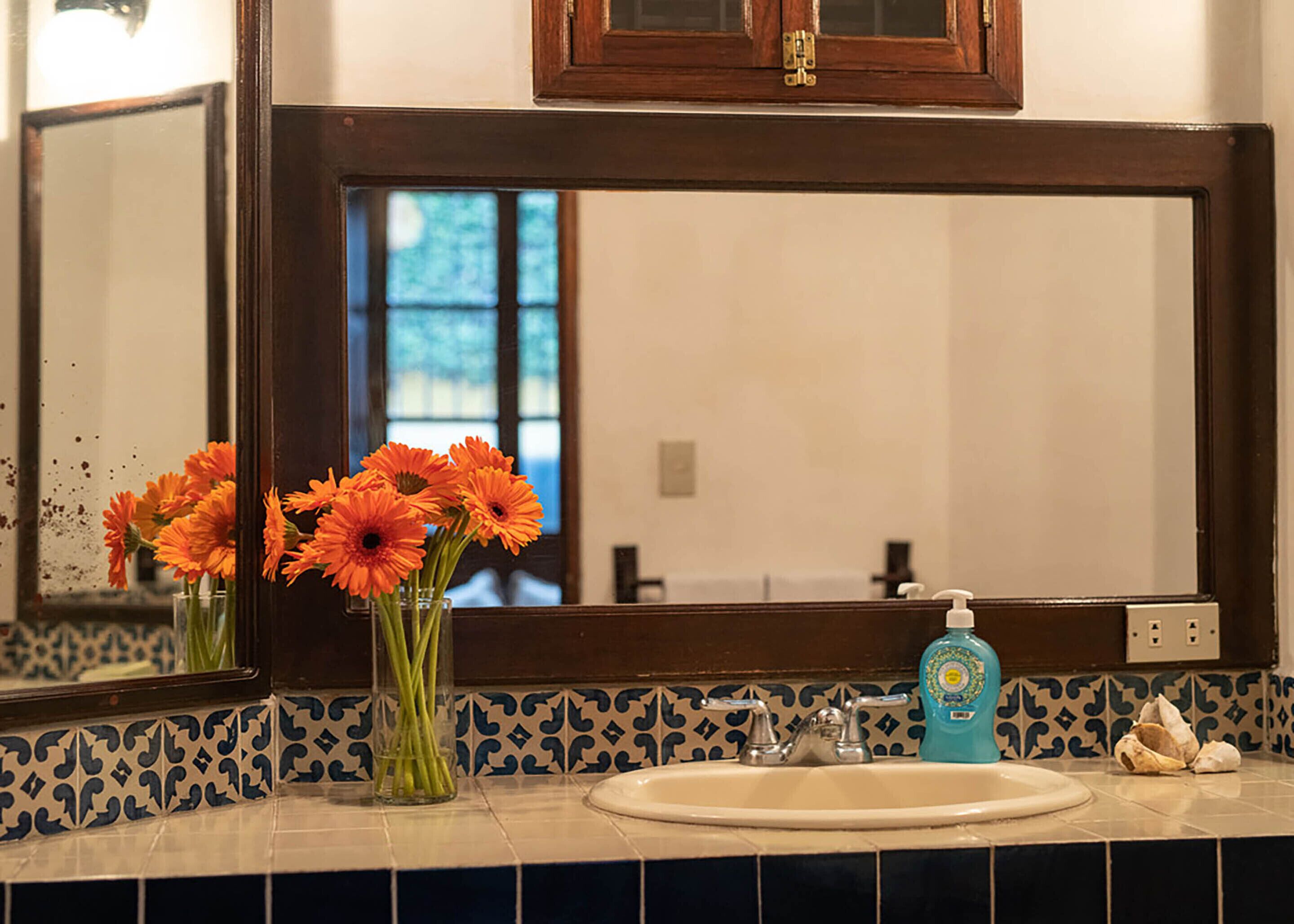 A bathroom with a large mirror, a sink, and a vase of orange flowers.