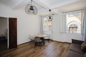 A room with a wooden floor, a round table with chairs, a brown sofa, and a large window with shutters. - Alloro Apartment - Three Bedroom Apartment, Sleeps 6 (Firenze)