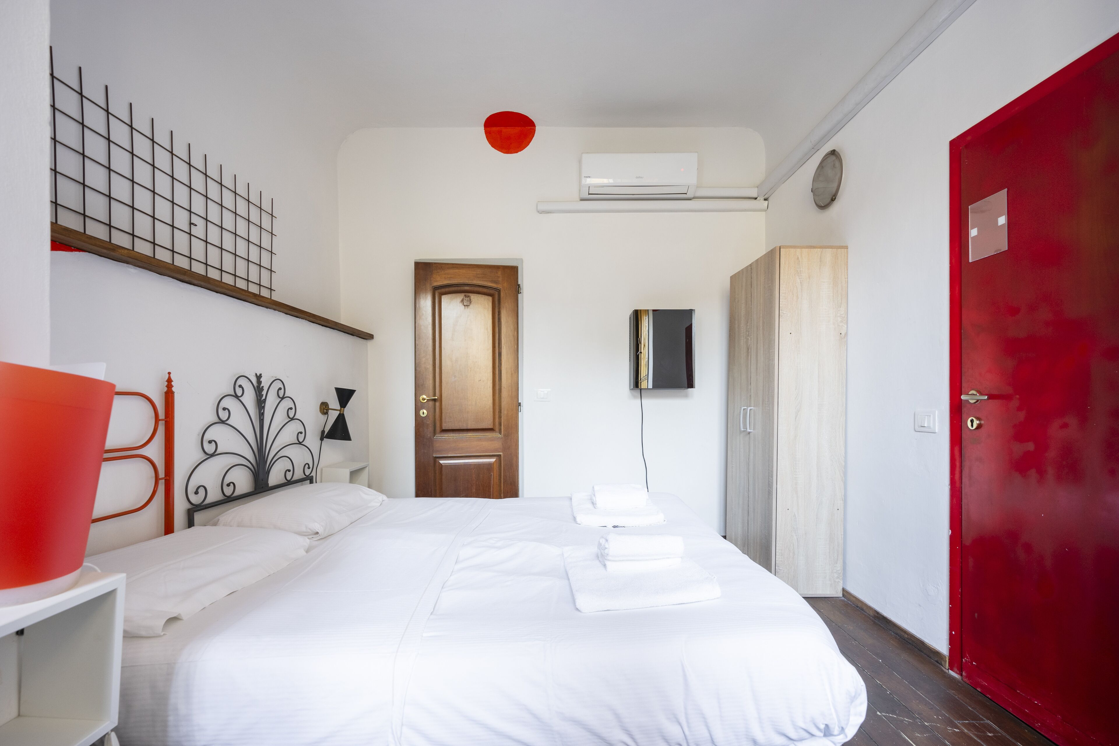 A hotel room with a bed, a red door, a wooden wardrobe, a white wall, and a red light fixture.