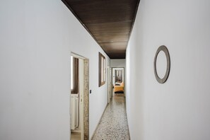 Ein Flur mit Holzboden, weißen Wänden und Fliesenboden. Am Ende des Ganges ist ein Bett mit gelber Decke zu sehen.