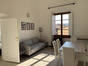 Une pièce comprenant un canapé, une table à manger avec des chaises, une lampe et une fenêtre donnant sur des immeubles.