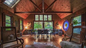 A rustic cabin interior with wooden walls, a dining area with a table and chairs, a large window with a view of greenery, and a chandelier. - Secluded in the Woods, Hot Tub (Twin Lakes)
