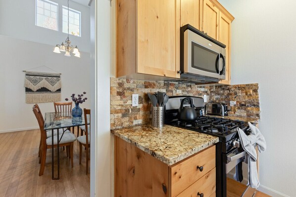 Una cocina con armarios de madera, encimera de granito, estufa y microondas. Hay una zona de comedor con una mesa y sillas, y un adorno decorativo en la pared.