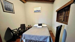 Ein kleiner, schlichter Raum mit einem Bett, einem Schreibtisch und einem Stuhl. Es gibt ein Fenster mit Gitter und ein Bild an der Wand.