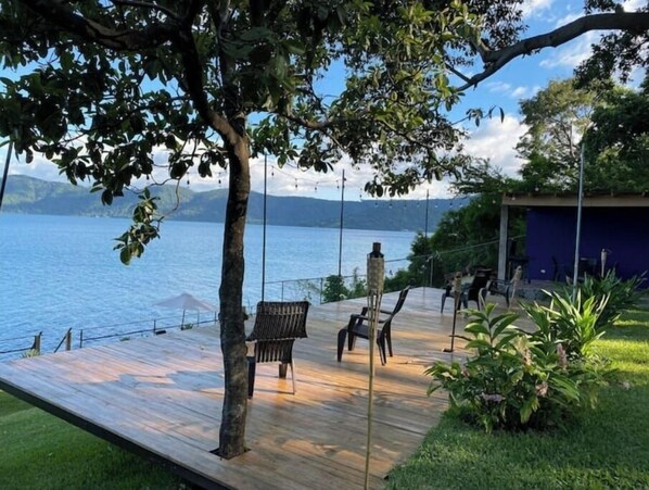Relax on the serene deck overlooking the lake, surrounded by lush greenery and distant mountains.