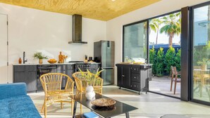 Eine moderne Küche mit einer großen Kochinsel, ein Essbereich mit Korbstühlen und eine Glastür, die auf eine Terrasse mit Blick auf Palmen führt.