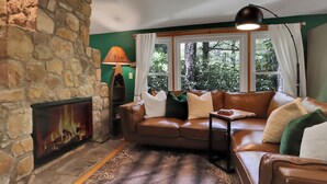 A cozy living room with a stone fireplace, a leather sofa, and a view of trees through large windows. - Lake, Golf & Hike at Linville Lodge in  Linville  Land  Harbor  Resort Community (Newland)