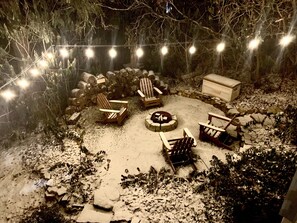 Un espacio al aire libre cubierto de nieve con sillas Adirondack y un brasero, rodeado de muros de piedra y luces colgantes.