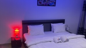 A neatly made bed with white linens and pillows, a bedside table with a lamp, and a framed picture on the wall. - LOYALTY HOTEL  (Accra)