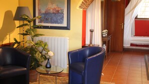 Una habitación con un sofá de cuero azul, una mesa de centro de cristal, una planta en maceta, un cuadro enmarcado y una cama con pared roja y cortinas blancas.