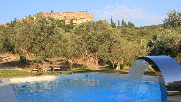 Una piscina con fuente acuática, rodeada de árboles y un edificio al fondo.