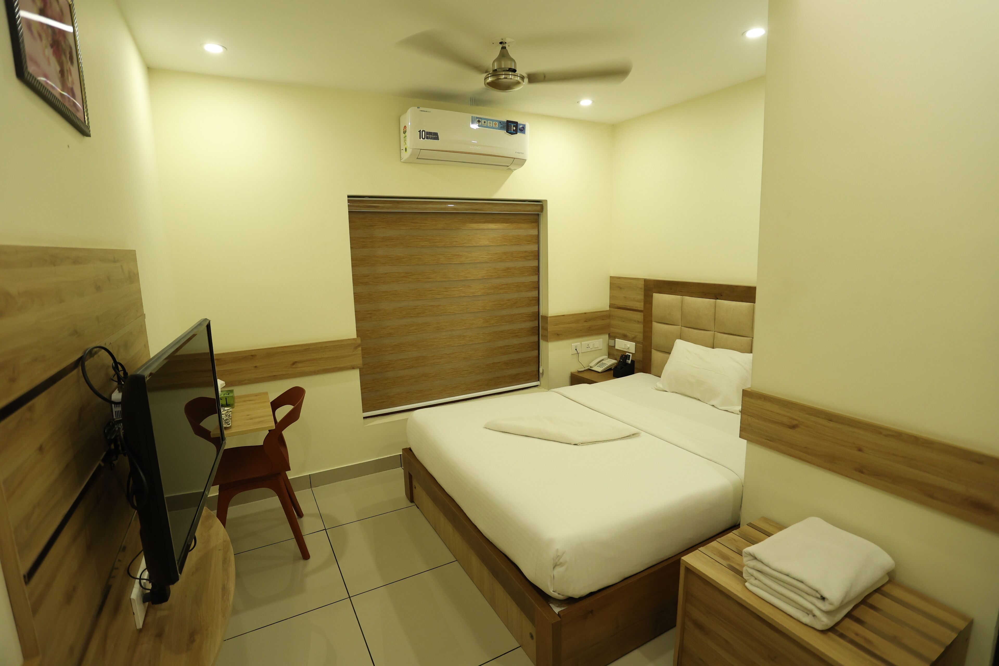 A hotel room with a bed, bedside table, a TV mounted on the wall, a window with blinds, and an air conditioning unit on the ceiling.