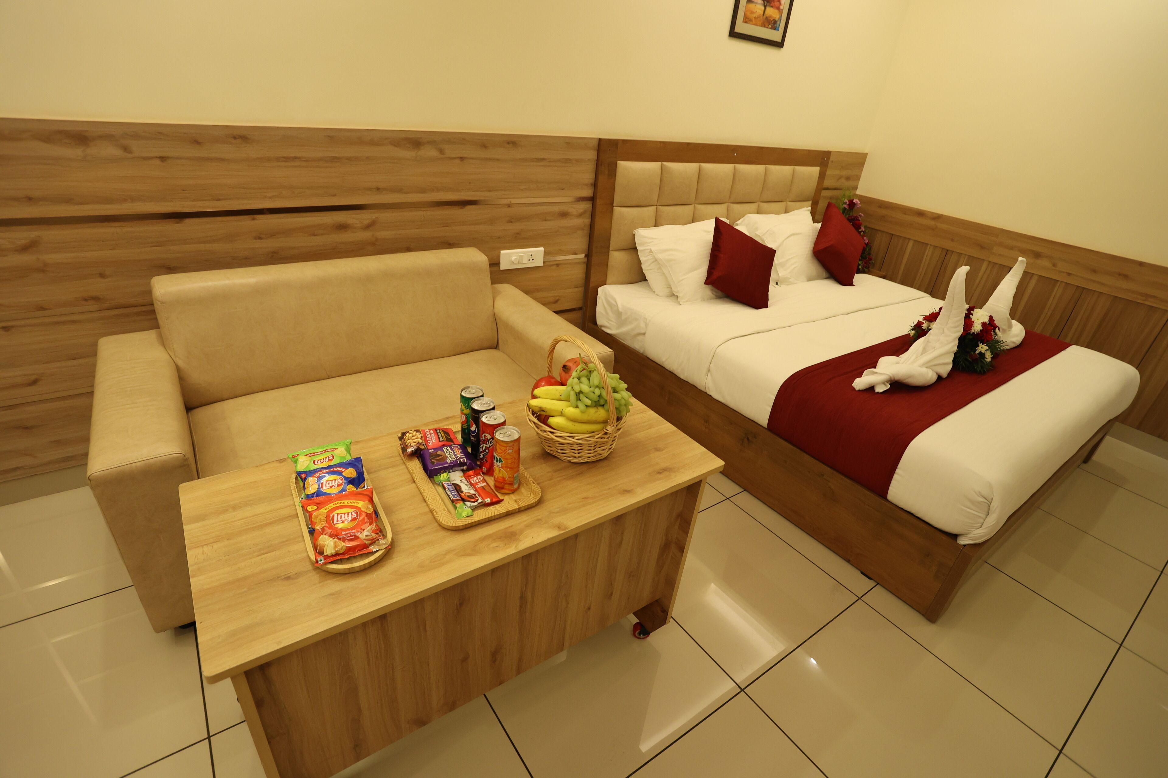 A hotel room with a sofa, a bed with a red blanket, and a table with snacks and fruits.