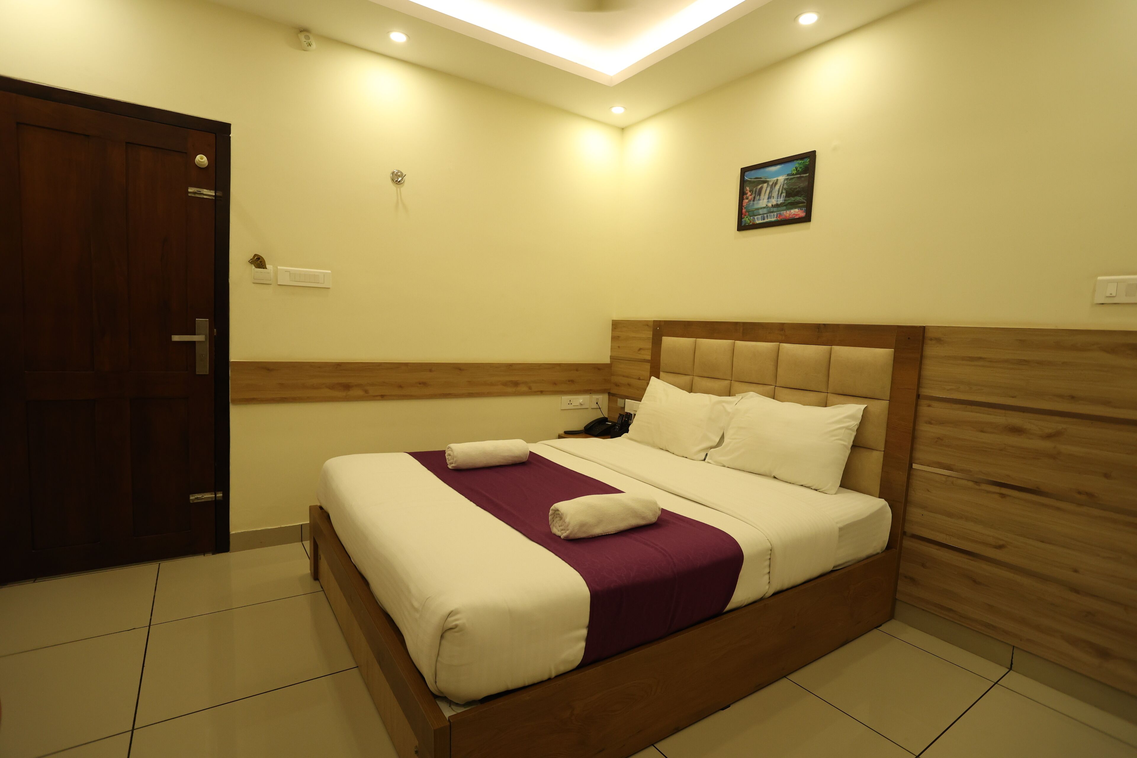 A neatly made bed with white and purple bedding, a wooden headboard, and a small wall-mounted picture.