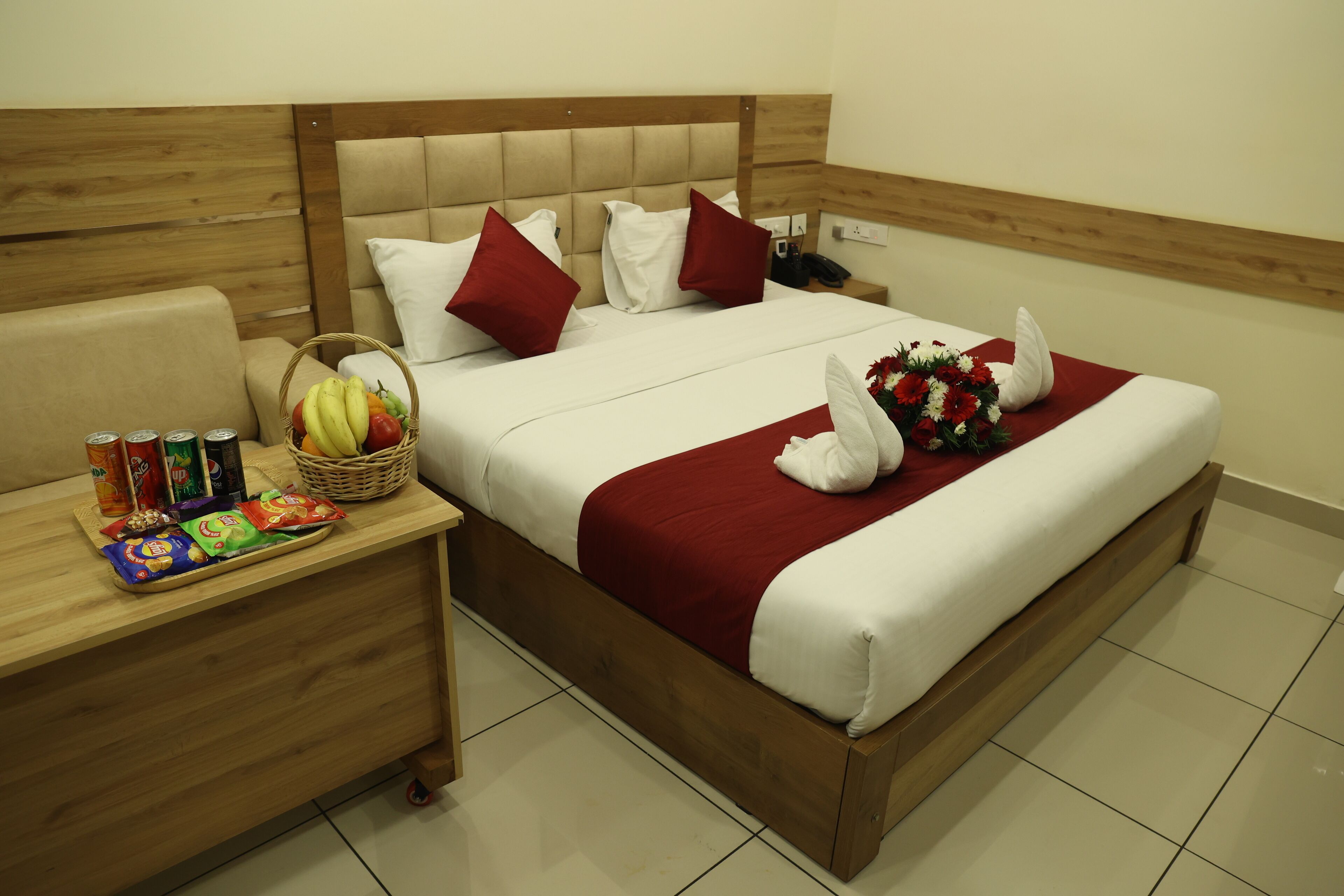 Quarto de hotel com cabeceira de madeira, cama bem arrumada com roupa de cama vermelha e branca, mesa de cabeceira com lanches e uma cesta de frutas, e manta vermelha.