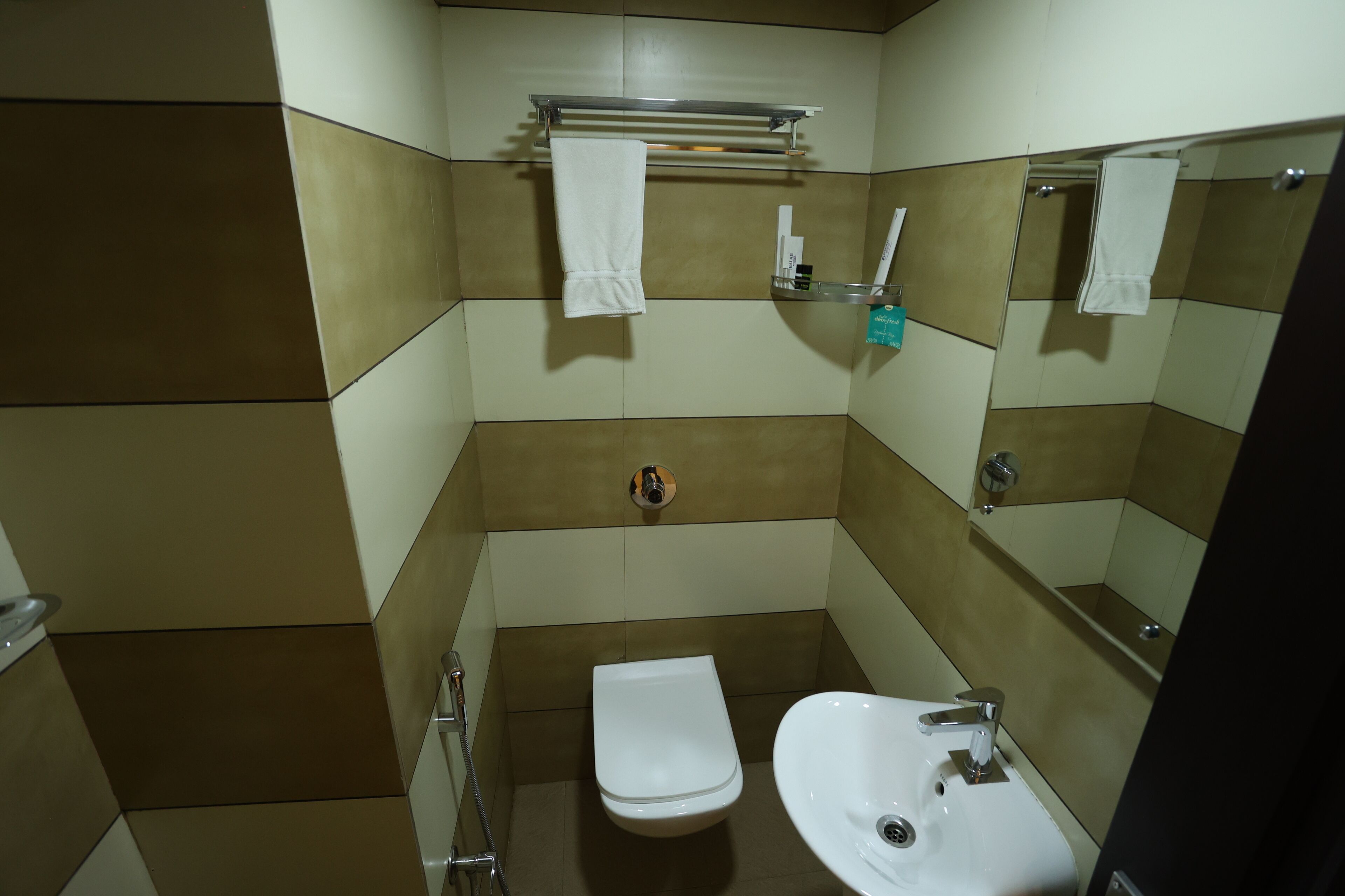 Un baño pequeño con inodoro, lavamanos y espejo. Hay un perchero sobre el inodoro y una repisa con artículos de aseo.