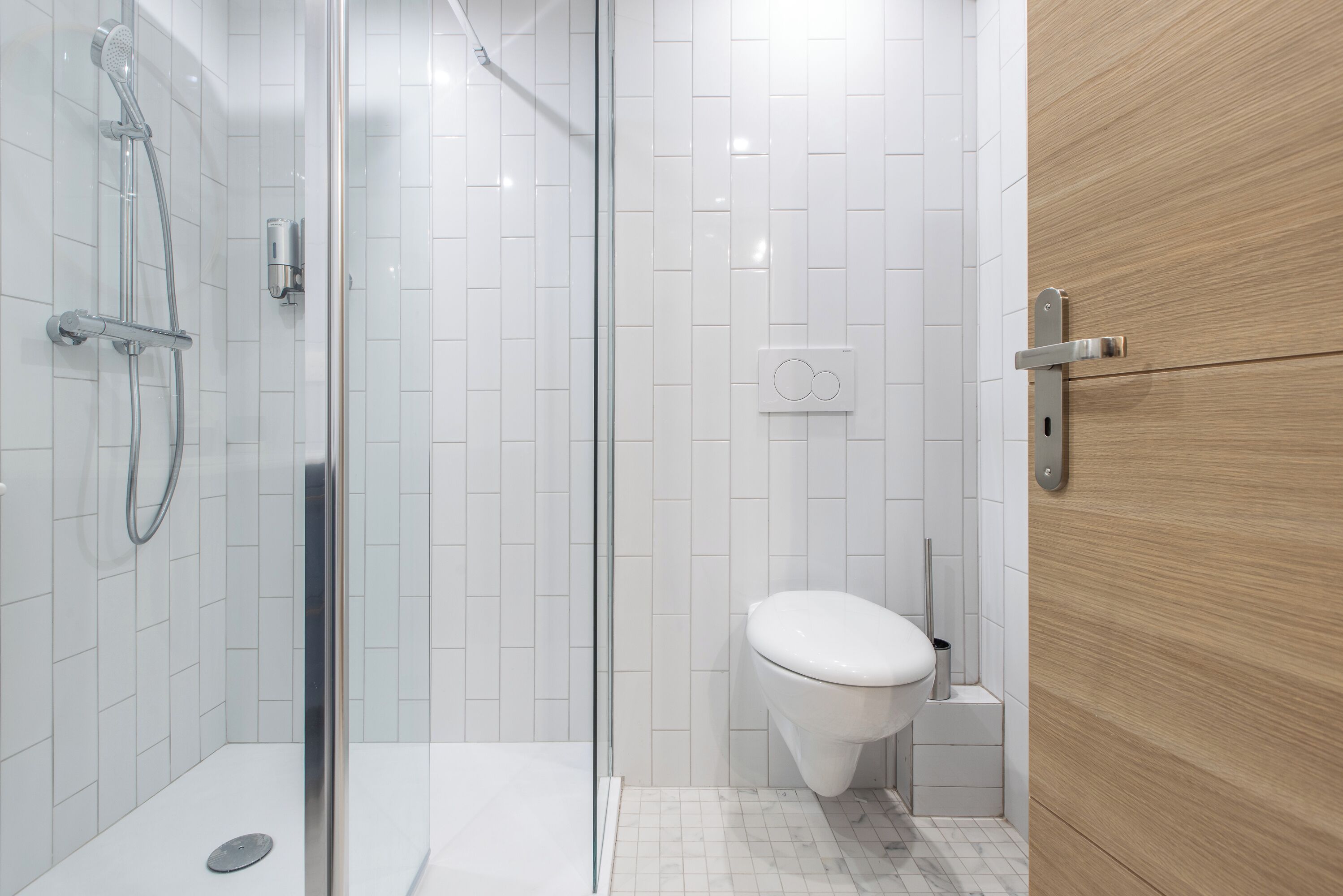 Et moderne badeværelse med et glasafskærmet brusebad, hvidmalede vægge, et hvidt toilet og en trædør.
