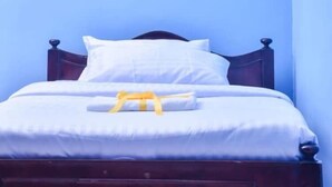 A bed with white bedding and a yellow object on it, a wooden headboard, and a blue wall. - Rovers Place Hotel  (Ahero)
