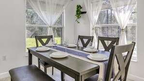Une table à manger dressée pour quatre personnes, avec de l’argenterie blanche, des verres à vin, des chaises en bois foncé et de fines rideaux blancs.