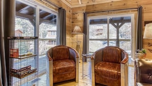 A cozy room with wooden walls and large windows, featuring two leather armchairs and a bookshelf with board games.