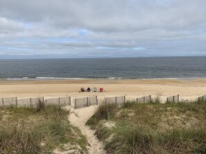 Uma praia com um caminho de areia que leva a cadeiras de praia e uma cerca de madeira.