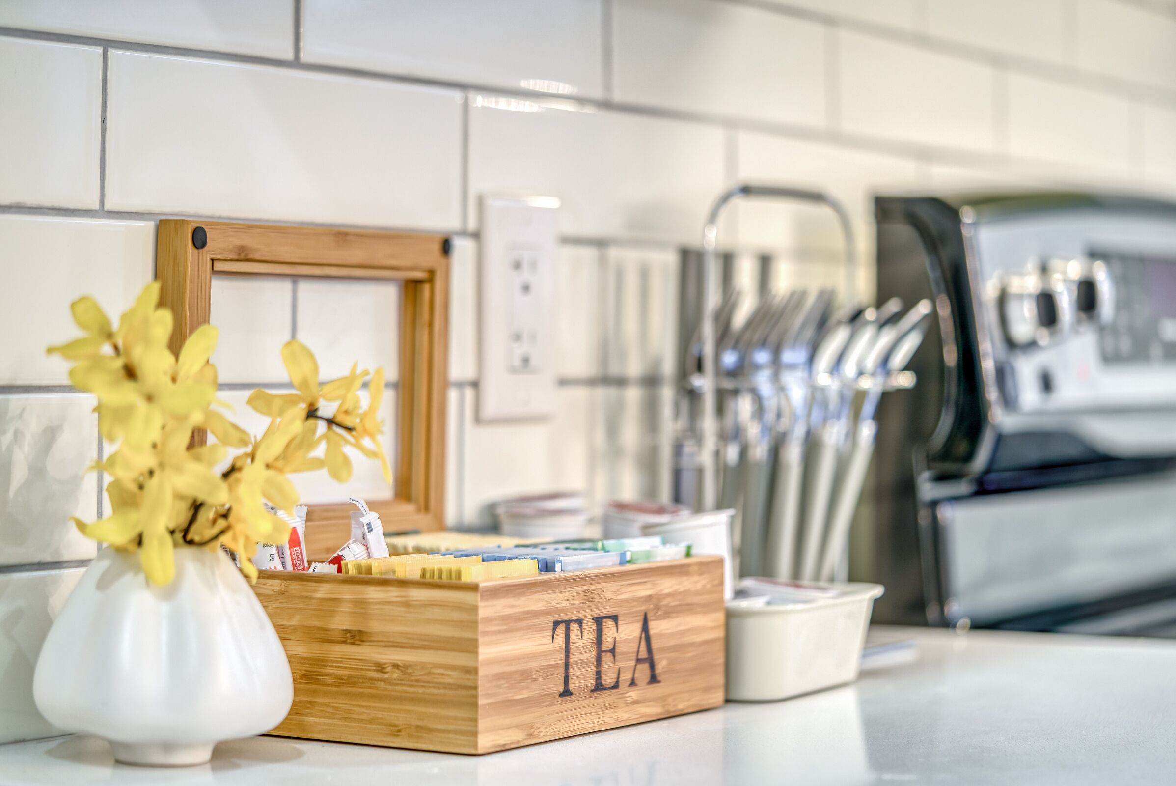 A kitchen counter with a wooden box labeled "TEA" containing tea bags and a white vase with yellow flowers.