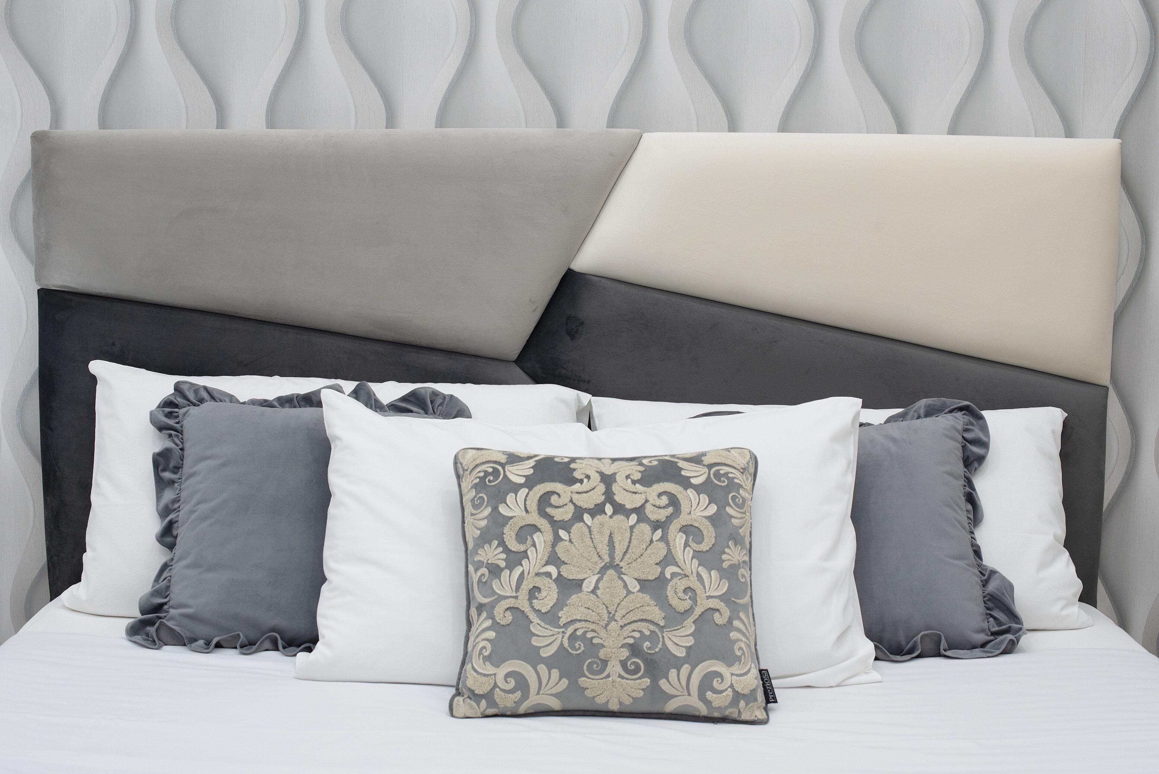 A bed with a headboard featuring a geometric pattern and three pillows in varying shades of gray and white.
