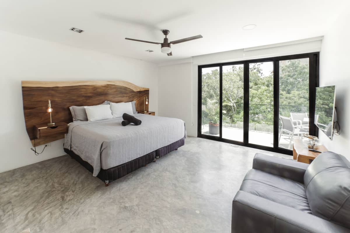 A modern bedroom with a large bed, a ceiling fan, and a view of greenery through large windows.