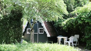 Ein kleines, dunkelgrünes Haus mit geneigtem Dach, umgeben von üppigem Grün und einem Tisch mit Stühlen.